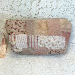 May include: A patchwork cosmetic bag with a beige zipper and a tassel. The bag is made of various fabric patches in shades of pink, beige, and cream, with floral and striped patterns. The bag is approximately 8 inches long.