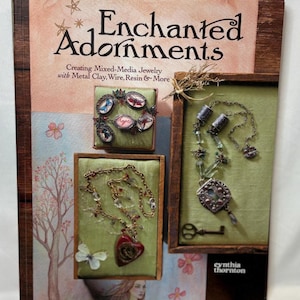 May include: A book cover titled "Enchanted Adornments" with jewelry examples. The cover features framed jewelry pieces with a heart pendant, a key, and a dragonfly design. The book's subtitle reads "Creating Mixed-Media Jewelry with Metal Clay, Wire, Resin & More."