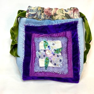 May include: A handmade patchwork bag with a light blue denim base. The bag features a square of purple velvet, lace, and floral embellishments. Green velvet straps and a floral patterned top complete the design.