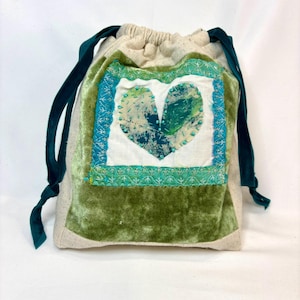 May include: A drawstring bag with a heart design. The bag features a cream-colored fabric body with a green velvet base. A square patch with a heart made of green and blue fabric is centered on the front. Dark green velvet ribbons serve as drawstrings.
