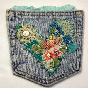 May include: A denim pocket, embellished with a heart-shaped design. The heart is composed of fabric patches, lace, beads, and sequins in shades of teal, pink, and blue. A button is centered on the left side. The top edge has a ruffled, light blue trim.