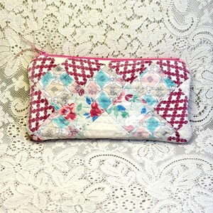 May include: A rectangular quilted zipper pouch featuring a patchwork design of white, pink, and teal fabric squares. The pattern includes floral motifs and a repeating dark pink lattice print, finished with a matching pink zipper closure.