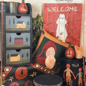 May include: A collection of decorative items including a black three-drawer chest with house paintings, a red wooden welcome sign with a rabbit, a black bucket labeled Trick with two figures, a round black box, and several orange gourd-like faces.
