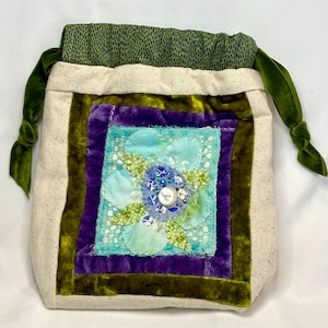 May include: A handmade drawstring bag with a floral design. The bag features a central square with a light blue flower, surrounded by purple and green velvet borders. The bag's body is a natural linen color, with green velvet drawstrings.