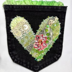 May include: Black denim pocket embellished with a heart-shaped design. The heart is crafted from lace, fabric scraps, and beads in shades of green, white, and red. Two white buttons and a green ruffled trim complete the design.