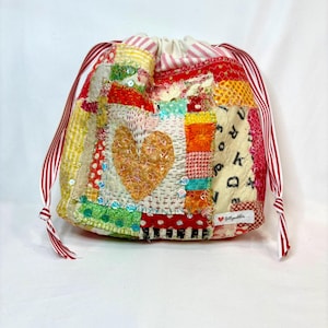 May include: A patchwork drawstring bag with a heart applique. The bag features a variety of colorful fabric scraps, including red, yellow, and green. The drawstring is red and white striped. The bag has a small label that says "Jellypuddler".