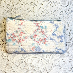 May include: A rectangular quilted zipper pouch featuring a patchwork design of white fabric with small floral patterns in shades of pink, light blue, and green. The pouch has a horizontal zipper closure along the top edge and visible stitching.
