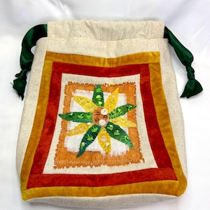 May include: A beige drawstring bag with a colorful floral design. The bag features a square patchwork design with a yellow, orange, and red border. The center has a white square with a flower made of green and yellow petals. Green satin ribbons serve as drawstrings.