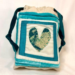May include: A handmade drawstring bag with a teal and white patchwork design. The bag features a teal velvet trim and teal drawstrings. A heart-shaped appliqué is centered on the front.
