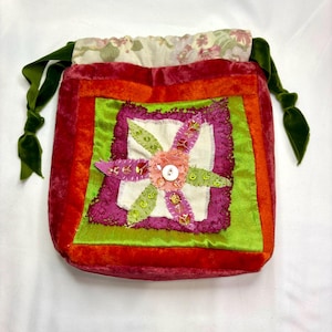 May include: A handmade drawstring bag with a patchwork design. The bag features a vibrant color palette of red, orange, green, and purple velvet. A floral applique with a button center adorns the front. Green velvet ribbons serve as drawstrings.