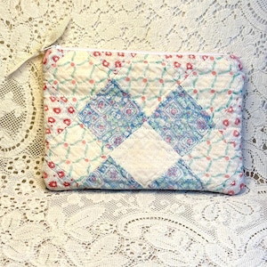 May include: A rectangular quilted zipper pouch featuring a patchwork design of white fabric with red floral patterns, light blue floral squares, and a white grid pattern with small pink dots. The pouch includes a white zipper and a beige fabric pull.