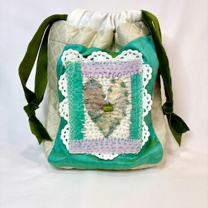 May include: A handmade drawstring bag with a quilted cream-colored body and a teal front panel. The front features a heart-shaped patch with green and purple accents, framed by white lace and green stitching. Green velvet ribbons serve as drawstrings.
