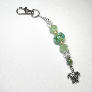 May include: A beaded keychain featuring a silver-toned metal lobster clasp attached to a series of light green, clear, and patterned glass beads. The beads are arranged in a vertical line, ending with a small, detailed silver-toned sea turtle charm.