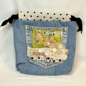 May include: A denim drawstring bag with a decorative pocket featuring floral embellishments, sequins, and buttons. The bag's top is cream-colored with black polka dots, and it has black ribbon drawstrings.