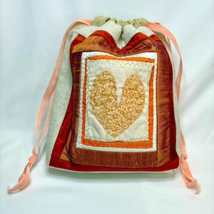 May include: A drawstring pouch with a heart design. The pouch features a central square with a gold heart on a cream background, framed by orange and red fabrics. Pink ribbons serve as drawstrings.