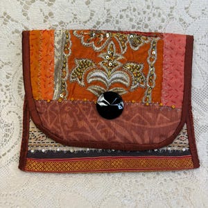 May include: A handmade clutch with a flap closure. The clutch features a patchwork design with orange, brown, and gold fabrics. The flap is adorned with sequins and a large black button. The overall style is bohemian.