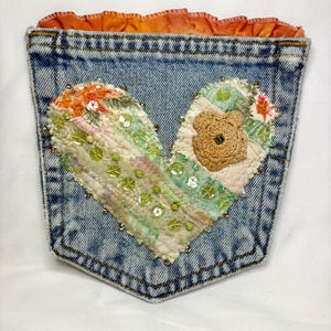 May include: A denim pocket with a heart-shaped patchwork design. The heart is made of fabric scraps in pastel colors, embellished with sequins, beads, and a button. The pocket's top edge has an orange ruffled trim.