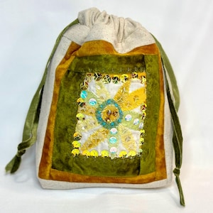 May include: A drawstring bag with a sunflower design. The bag features a cream-colored body with a green velvet panel and a gold border. The sunflower is made of yellow fabric and embellished with sequins. Green velvet ribbons serve as drawstrings.