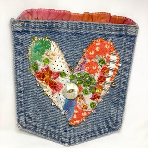 May include: A denim pocket with a heart-shaped appliqué. The heart is made of colorful fabric scraps, sequins, and beads, with a mother-of-pearl button in the center. The pocket's top edge has a ruffled pink and orange fabric.