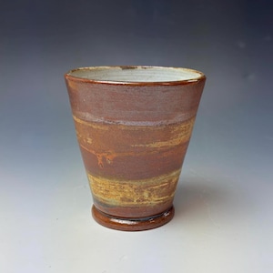 May include: A handmade ceramic tumbler with a brown and yellow striped design. The cup has a wide mouth and a small, rounded base. The interior is a light cream color. The tumbler is suitable for hot or cold beverages.