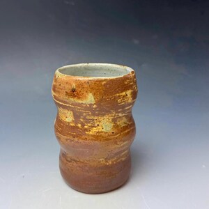 May include: A handmade ceramic tumbler with a unique, curved shape. The exterior features a blend of brown and tan hues, with a speckled texture. The interior is a light, speckled color.
