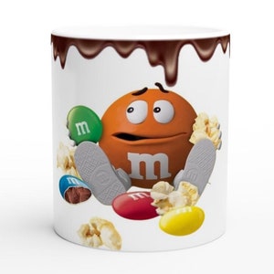 Puede incluir: Taza de cerámica blanca con un glaseado de chocolate marrón que gotea por el lado. La taza presenta un personaje M&M naranja con una expresión de sorpresa, rodeado de caramelos M&M verdes, rojos, amarillos y marrones y palomitas de maíz.