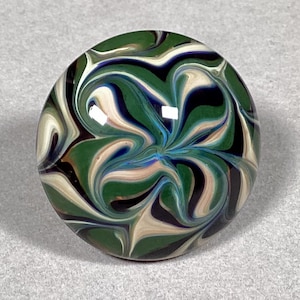 May include: A round, glass marble with a swirling pattern of green, black, white, blue, and tan colors. The marble has a glossy finish and a smooth surface. The design resembles a flower or starburst.