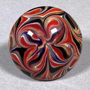 May include: A round, glass marble with a swirling pattern of red, black, white, blue, and brown colors. The marble has a glossy finish and a central vortex design.