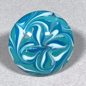 May include: A round, glass marble with a swirling pattern of teal, white, and various shades of blue. The marble has a glossy finish and a smooth surface. The design is reminiscent of ocean waves.