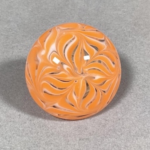 May include: A glass marble with a swirling orange and white pattern. The marble is round and smooth.