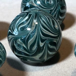 May include: A set of three glass marbles with a swirling teal and white pattern. The marbles are shiny and have a speckled effect.