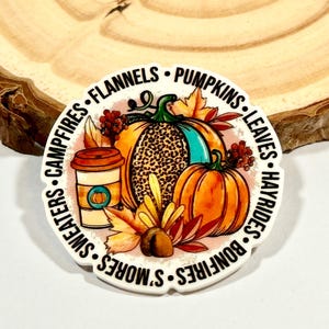 May include: A round sticker with a fall theme. It features pumpkins, leaves, a cup of coffee, and the text "S'mores, Bonfires, Hayrides, Leaves, Pumpkins, Flannels, Campfires, Sweaters".