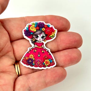 May include: A die-cut sticker featuring a cartoon character with large black eyes and a sugar skull face design. The character wears a vibrant red dress adorned with colorful floral patterns and a large headpiece of bright red, yellow, and purple flowers.