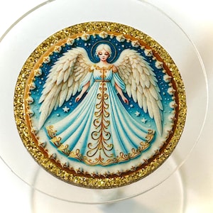 May include: Round angel ornament with a gold glitter border. The angel has white wings, a blue dress, and a halo. The background is blue with white stars. The ornament is on a clear, round surface.