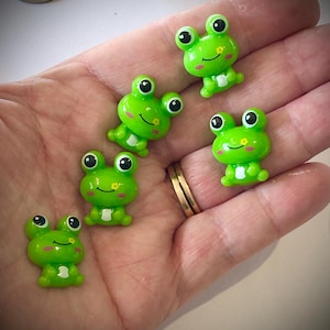 May include: Five small, bright green frog figurines with large black and white eyes and smiling faces. The frogs have white bellies and are posed in various positions. The figurines are held in a person's hand.