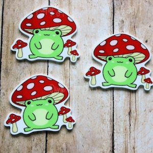 May include: Three green frog stickers with red and white mushroom hats. Each frog is sitting on a mushroom with white spots. The mushrooms are red with white spots.