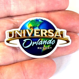 May include: A circular sticker featuring the Universal Orlando Resort logo. The design includes a blue and green globe graphic with the word UNIVERSAL in gold metallic-style lettering and the word Orlando in blue script, with RESORT below.