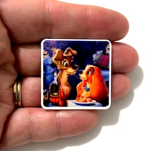 May include: A square sticker featuring a scene of two dogs, one tan and one brown with long ears, sharing a plate of spaghetti. The background shows a dimly lit restaurant setting with a candle and a musician playing an accordion in the shadows.