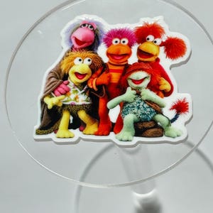 May include: A flat, die-cut acrylic charm featuring a colorful group of five Fraggle Rock characters. The characters include a purple-haired figure, a yellow-furred figure in a floral shirt, a bright orange figure, a red-haired figure, and a green figure.