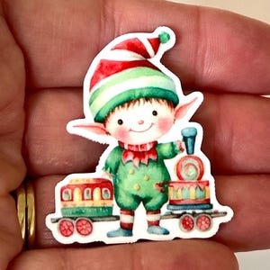 May include: A sticker of a cheerful elf in a green outfit and a red and green striped hat. The elf is holding a blue train engine and standing next to a red, green, and yellow train car. The sticker has a white border.