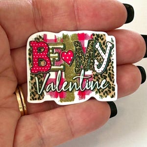 May include: A rectangular Valentine's Day themed sticker with the words "BE MY Valentine". The text is in a mix of fonts and colors, including red, white, and black. The background features a plaid pattern and leopard print.
