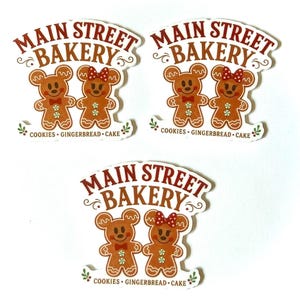 May include: Three die-cut stickers featuring the text MAIN STREET BAKERY in brown letters. Below the text are two brown gingerbread bear characters, one with a red bow. The bottom text reads COOKIES GINGERBREAD CAKE with small green holly accents.