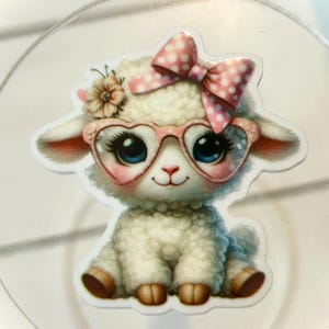 May include: A cute cartoon lamb sticker with a pink bow, heart-shaped glasses, and a flower. The lamb has big blue eyes and is sitting down. The sticker is on a clear, round surface.