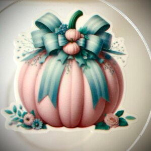 May include: A die-cut sticker featuring a soft pink pumpkin adorned with a large, striped teal ribbon bow at the top. The pumpkin is decorated with small pink and teal floral accents at the base and around the stem, creating a festive appearance.