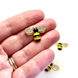 May include: A gold-toned bee brooch with black and yellow stripes on the body and crystal-studded wings. The bee has a black head and is set against a white background. The brooch is a jewelry item.