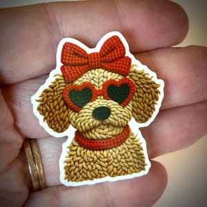 May include: A sticker featuring a cartoon dog with a knitted appearance. The dog wears heart-shaped sunglasses, a bow, and a collar, all in shades of red. The dog's fur is a mix of tan and brown.