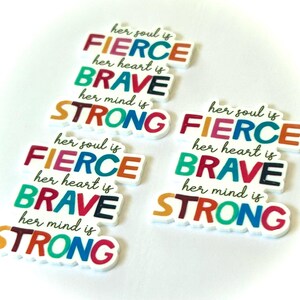 May include: Several white, rectangular charms with the words "her soul is FIERCE, her heart is BRAVE, her mind is STRONG" in colorful block letters. The charms are in various colors including orange, green, red, and blue.
