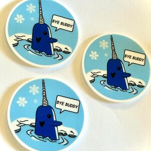 May include: Three round stickers featuring a blue narwhal with a spiraled horn, swimming in icy water. Each sticker has a speech bubble with the text "BYE BUDDY" and snowflakes on a light blue background.