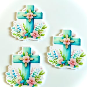 May include: Three turquoise wooden crosses with floral embellishments. Each cross is surrounded by pink and white flowers, green leaves, and blue accents.