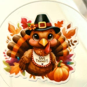 May include: A cartoon turkey wearing a pilgrim hat and a bib that says "Give Thanks". The turkey is surrounded by fall leaves and a pumpkin.